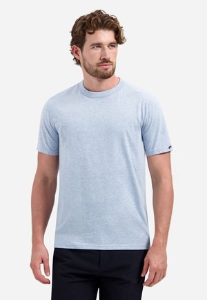 Man met kort krullend haar, gekleed in een lichtblauw T-shirt met korte mouwen en ronde hals, en donkerachtige broek, staand tegen een witte achtergrond.