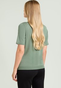 Top de manga corta y ajustado en tejido de punto suave de color verde claro. Cuenta con escote redondo y dobladillo acanalado. Combinado con pantalones negros, mostrado desde la parte trasera.