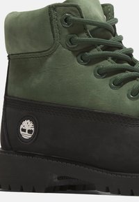 Stivale in pelle verde con finitura testurizzata, accenti neri, lacci tondi e una toppa a logo circolare sul lato. Suola in gomma spessa.