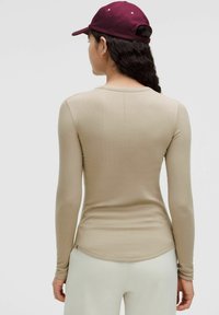 Camicia aderente a maniche lunghe color beige, che crea una silhouette morbida, caratterizzata da un collo rotondo e un orlo leggermente curvo. Il retro presenta un dettaglio di cucitura.