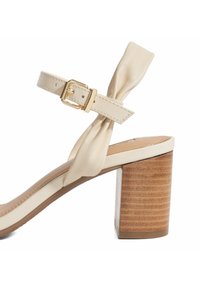 Beige heeled sandal with a strap secured by a gold buckle. Features a knotted design and a textured wooden block heel. Smooth leather texture.
