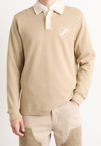 Beige långärmad polo med vit krage. Har en broderad sköldlogotyp på vänster bröst och en slät tygtextur.