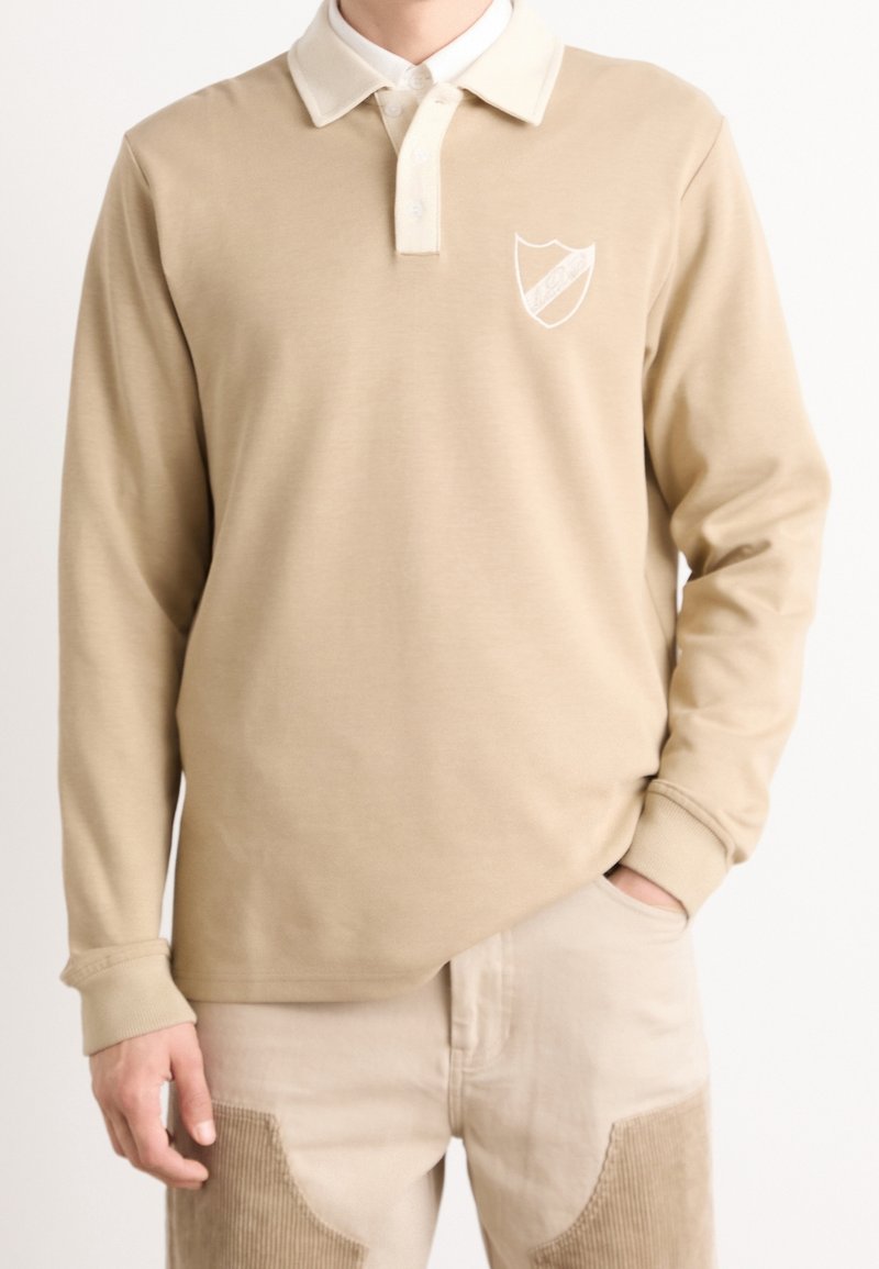 Beige långärmad polo med vit krage. Har en broderad sköldlogotyp på vänster bröst och en slät tygtextur.