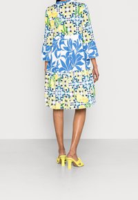 Vestido de estampado floral en azul, amarillo y verde, con mangas tres cuartos, un diseño en capas y un corte relajado. Sandalias de tacón alto amarillas.