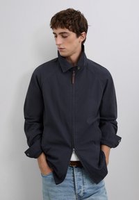 Veste légère marine avec un col haut, fermeture éclair à l'avant avec tirette en cuir et poignets boutonnés. Tissu lisse et coupe décontractée.