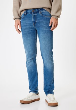 Vaqueros de mezclilla azul de corte slim, con cierre de botón, cinco bolsillos y un ligero desvanecimiento. Combinados con zapatillas blancas.