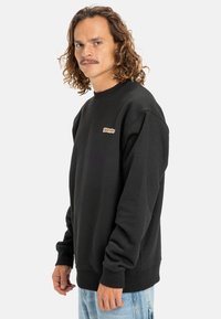 Sudadera de algodón negra con puños y cuello acanalados. Presenta un pequeño logo colorido en el pecho. El modelo se encuentra contra un fondo neutro.