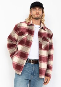 Camisa de franela en un patrón de cuadros en rojo, crema y beige. Presenta un cierre de botones en la parte delantera, dos bolsillos en el pecho y un corte holgado. Usada sobre una camisa blanca.