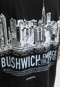 Fekete póló, fehér grafikával, ami egy város sziluettjét ábrázolja, vastag betűkkel "BUSHWICH" felirattal, alatta egy laza betűtípussal írt leíró szöveggel.