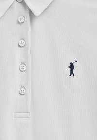 Vit polo skjorta i strukturerad tyg med en knapplist med tre knappar och en liten mörkblå broderad logotyp på bröstet.