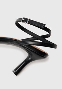 Musta nahkainen korkokenkä, jossa on ohut nilkkahihna ja hopeinen solki valkoisella taustalla.