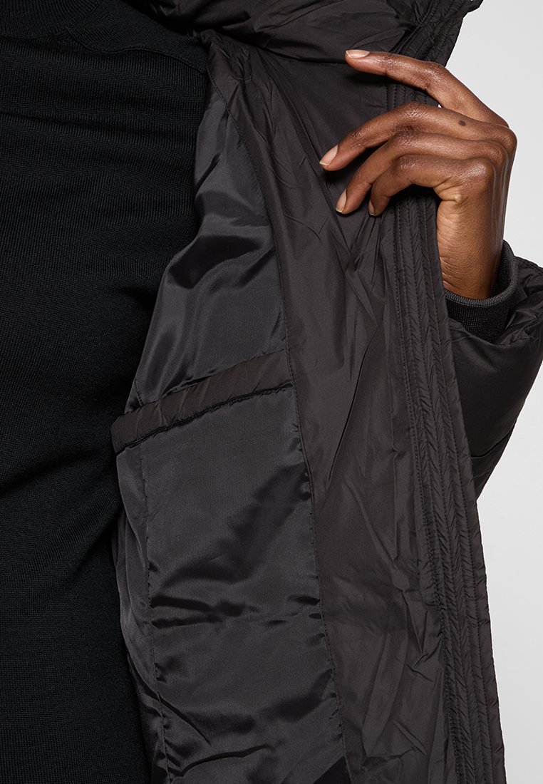 Veste noire isolée avec un extérieur lisse et brillant. Présente une poche intérieure zippée et des poignets côtelés pour plus de chaleur et un ajustement optimal.