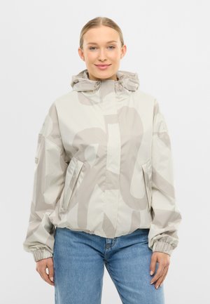 Uśmiechnięta kobieta ubrana w jasnobeżową, wzorzystą kurtkę z kapturem oraz niebieskie dżinsy, stojąca na jednolitym białym tle.