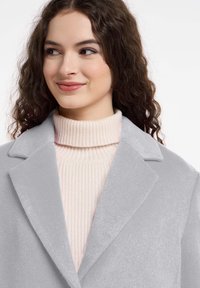 Grijze fleece jas met een brede kraag, gedragen over een lichtroze geribbelde coltrui. Heeft een zachte textuur en een strakke silhoutte.