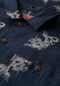 Camisa de manga corta azul marino hecha de tela con ilustraciones de dragones blancos. Incluye botones marrones y una pequeña etiqueta de marca.