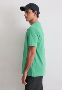 Zielona bawełniana koszulka z krótkimi rękawami i okrągłym dekoltem, zestawiona z ciemną czapką. Tkanina ma gładką fakturę i luźny krój.