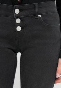 Μαύρο τζιν με ψηλή μέση, που διαθέτει τρία ασημένια κουμπιά στο μπροστινό μέρος, ελαφρώς ελαστικό ύφασμα και κλασικό σχέδιο με πέντε τσέπες.