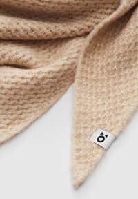 Écharpe tricotée douce et beige avec un motif texturé et une étiquette blanche affichant un logo circulaire noir avec deux petits triangles.