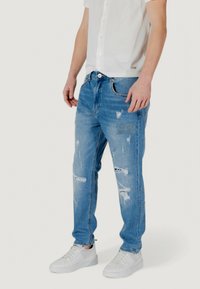 Jeans de color azul claro, desgastados, con múltiples desgastes y un parche. Cintura media con cierre de botón y cremallera. Combinados con zapatillas blancas.