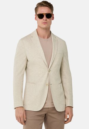 Blazer beige claro con textura, dos botones y bolsillos frontales, llevado sobre un suéter marrón claro, complementado con unas gafas de sol oscuras.