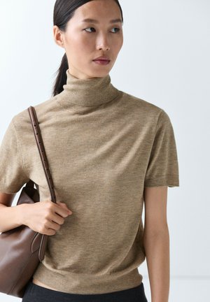 Femme portant un pull à col roulé à manches courtes beige, tenant un sac à bandoulière en cuir marron, regardant vers sa gauche sur un fond clair uni.