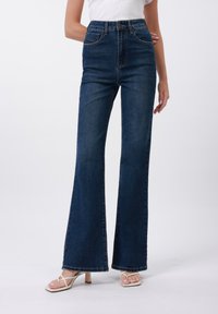 High-waisted flared jeans in dark blue denim with a smooth texture, featuring front pockets and a sewn hem line. Paired with white sandals.