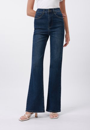Jeans a vita alta svasati in denim blu scuro con una texture liscia, dotati di tasche anteriori e una linea dell'orlo cucita. Abbinati a sandali bianchi.