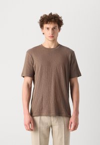 Brown short-sleeve t-shirt in a soft, textured fabric with a round neckline and subtle branding on the chest, paired with light-colored trousers.