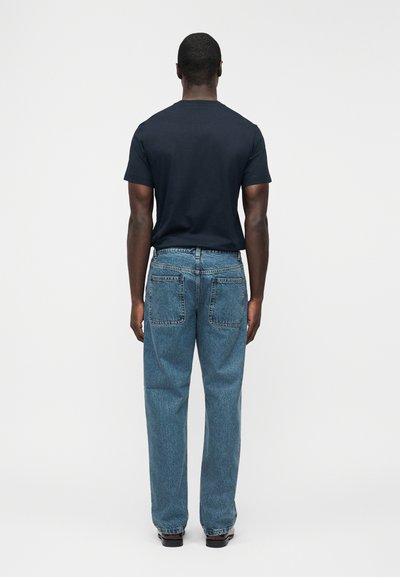 Άνδρας που στέκεται γυρισμένος με την πλάτη φορώντας μαύρο κοντομάνικο πουκάμισο, μπλε τζιν με πίσω τσέπες και σκούρα παπούτσια σε απλό λευκό φόντο.