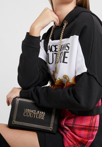 Persona che indossa un felpa con un motivo nero, bianco e rosso, mentre tiene una pochette nera di Versace Jeans Couture con dettagli in catena dorata.