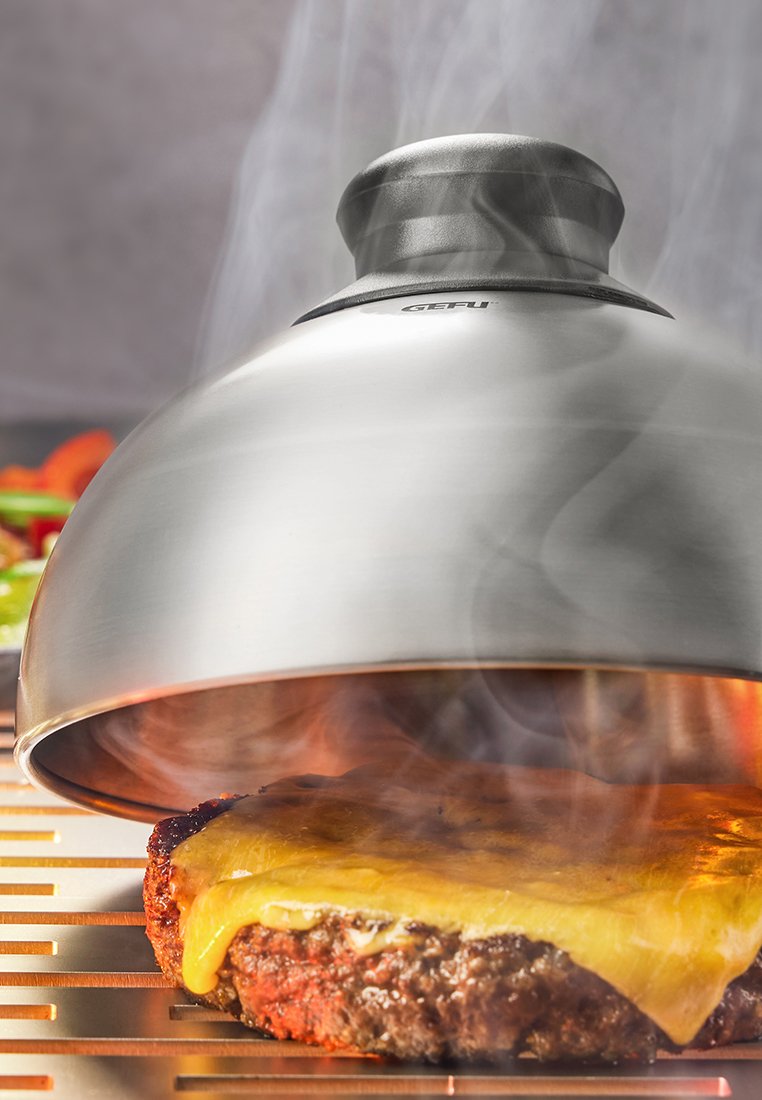 Stalowa pokrywa w kształcie kopuły na jedzenie z czarnym wierzchem, częściowo uniesiona, ukazująca kotlet wołowy z roztopionym żółtym serem.