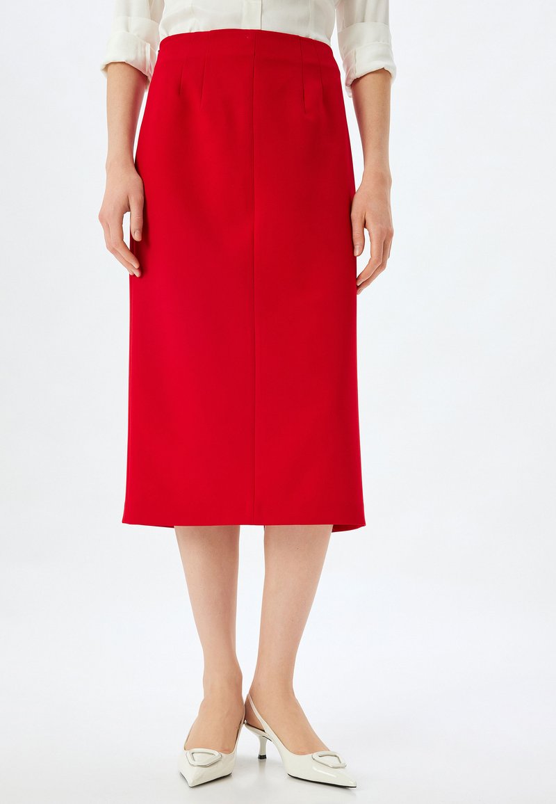 Roter Bleistiftrock mit klaren Linien, knielang, mit vorderen Falten. Kombiniert mit weißen Slingback-Absätzen, die ein quadratisches Detail zeigen.