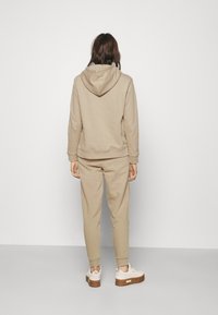 Žena stojící čelem ke zdi, oblečená v béžové mikině s kapucí, odpovídajícími tepláky a bílými teniskami s hnědými podrážkami na světlém šedém podlahovém povrchu.