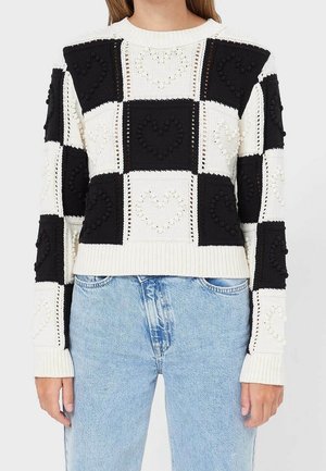 Femme portant un pull à carreaux noir et blanc avec des motifs en forme de cœur texturés, associé à un jean bleu clair.