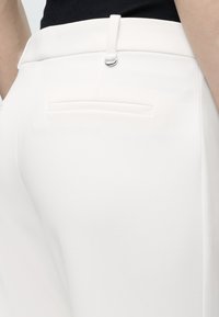 Κοντινό πλάνο ενός ατόμου που φοράει λευκό ραμμένο παντελόνι με πίσω τσέπη με μια επένδυση και λεπτομέρειες με θηλιές για ζώνη, συνδυασμένο με μαύρο τοπ.