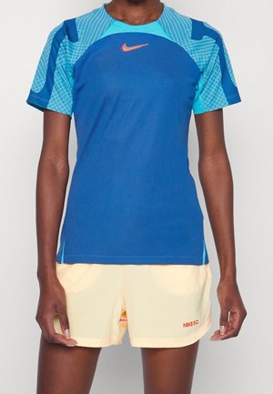 Person iført en blå og turkis kortærmet Nike-trøje og lysegule Nike F.C. shorts, der står mod en ensfarvet baggrund.