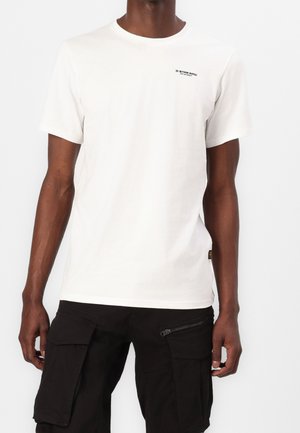 Camiseta blanca de algodón con cuello redondo, mangas cortas, discreto logo negro en el pecho y detalles en la etiqueta lateral. Combinada con pantalones cargo negros.