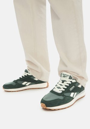 Green athletic sneakers featuring a mix of suede and mesh, with white accents and textured rubber soles, worn with light-colored pants.