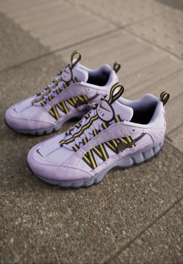 Baskets en daim violettes avec des lacets rayés jaunes et noirs, semelle en caoutchouc et un logo "AIR" bien en vue au niveau du talon, sur un fond texturé.