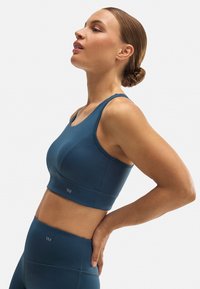 Sujetador deportivo en color verde azulado con escote redondo, diseño que ofrece soporte y textura sin costuras. Logo de la marca destacado en la parte inferior.