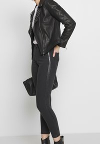 Chaqueta de moto de cuero negro con acentos de tachuelas, combinada con jeans negros ajustados que tienen una franja brillante en el lateral y botas de tobillo con tacón.
