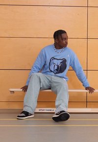 Világoskék pulóver medve grafikával, világoskék farmerrel és fekete-fehér tornacipővel párosítva. Egy fából készült padon ülve.