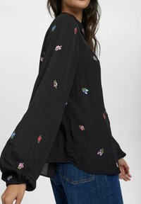 Schwarze Bluse mit buntem Blumenstickerei, langen Ärmeln und lockerer Passform. Der Stoff wirkt leicht und leicht strukturiert.