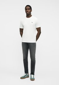 Άνδρας στέκεται με τα χέρια πίσω από την πλάτη, φοράει λευκό t-shirt, στενά σκούρα γκρι τζιν και πράσινα σουέντ αθλητικά παπούτσια με λευκά κορδόνια.