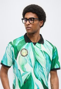 Άνδρας με γυαλιά που φοράει πράσινη ποδοσφαιρική φανέλα με φύλλωμα, μαύρο γιακά και σήμα ποδοσφαιρικής ομοσπονδίας του 1978.