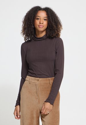 Femme aux cheveux bouclés portant un col roulé côtelé foncé et un pantalon en velours côtelé beige, debout devant un fond blanc uni.