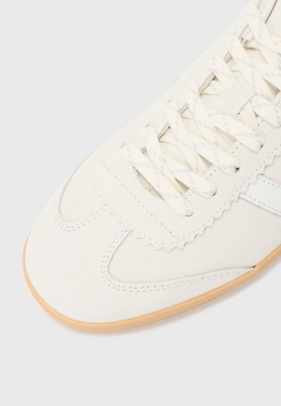 Chaussure de sport blanche en suede avec des lacets plats, un bord festonné et une semelle en caoutchouc beige. Présente une fine bande blanche subtile sur le dessus.