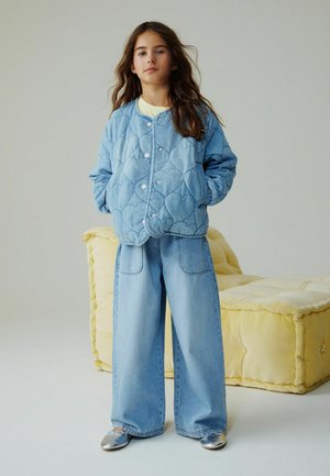 Mädchen mit langem Haar trägt eine blaue gesteppte Jacke mit Herzmuster, weit geschnittene Jeans und silberne Ballerinas, steht vor einem gelben gepolsterten Stuhl.