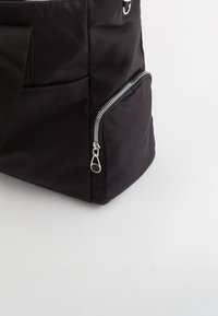 Bolso de mano negro de nailon con una superficie texturizada, que cuenta con un bolsillo lateral con cremallera y una correa ajustable. Diseño simple con acentos de hardware en plata.
