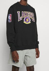 Sudadera negra con "LAKERS" en púrpura y amarillo, logo de la NBA en la manga, cuello redondo y puños y dobladillo acanalados.